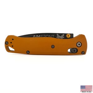 May include: A folding pocket knife with a textured orange handle and a black blade. The blade features the text "AIMERONT" and a butterfly logo. The handle includes a gold-colored thumb stud, several dark screws, and a lanyard hole at the end.