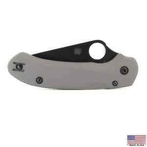 May include: A grey folding knife with a black blade. The knife has a textured handle and a circular opening near the blade's tip. The blade has a serrated edge near the handle. The knife has a small spider logo on the blade. Made in USA.