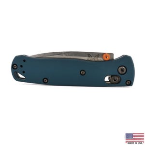 May include: A folding pocket knife with a matte teal handle and a partially visible grey blade. The handle features black screws, a lanyard hole, and a bright orange thumb stud. The Benchmade butterfly logo is etched on the blade surface.