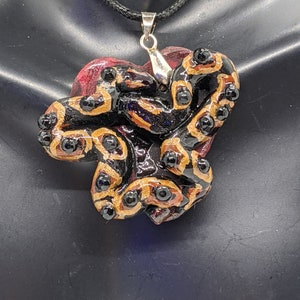May include: A black and gold snake pendant necklace with a red heart-shaped base. The snake is curled up in a heart shape.