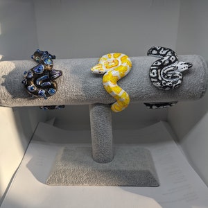 May include: Three snake-shaped bracelets on a gray display stand. The bracelets are made of black and white beads, yellow and white beads, and blue and black beads.