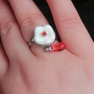 May include: A silver ring with a white flower design and a red glass bead accent.