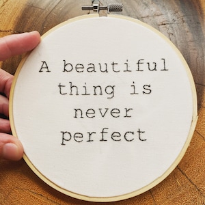 May include: A white embroidery hoop with the quote "A beautiful thing is never perfect" stitched in black thread.
