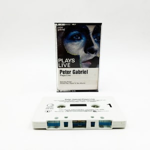 May include: A white cassette tape with the text "Peter Gabriel Plays Live" printed on the label. The cassette tape is in a clear plastic case with a black border. The case has a photo of Peter Gabriel on the front with the text "Plays Live" and "Peter Gabriel Plays Live" printed on it.