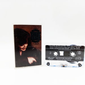 May include: A black cassette tape with a white label. The label has the text "Bonnie Raitt" and "Luck of the Draw" on it. The cassette tape is in a clear plastic case.