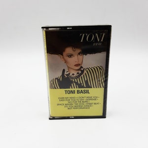Puede incluir: Una caja de cassette amarilla y negra con una foto de Toni Basil en la parte delantera. El texto en la caja dice "Toni Basil" y enumera las canciones "Over My Head", "I Don't Hear You", "Easy For You To Say", "Suspense", "Go For The Burn", "Space Walkin' The Dog", "Street Beat", "Do You Wanna Dance" y "Best Performance".