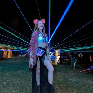 K&ouml;nnte beinhalten: Eine Frau tr&auml;gt einen schwarzen Rock mit einem durchsichtigen &Uuml;berzug, ein rosa-wei&szlig;es Crop-Top und eine holografische Bauchtasche. Sie steht vor einer B&uuml;hne mit Laserlichtern.