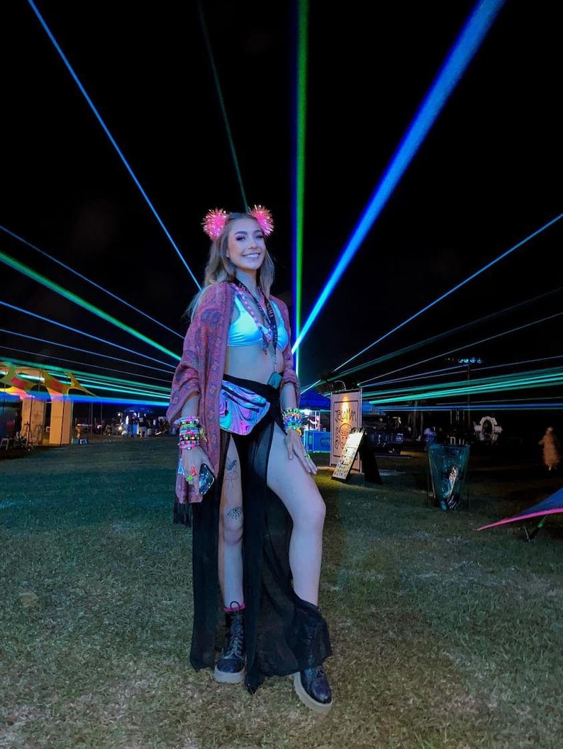 K&ouml;nnte beinhalten: Eine Frau tr&auml;gt einen schwarzen Rock mit einem durchsichtigen &Uuml;berzug, ein rosa-wei&szlig;es Crop-Top und eine holografische Bauchtasche. Sie steht vor einer B&uuml;hne mit Laserlichtern.