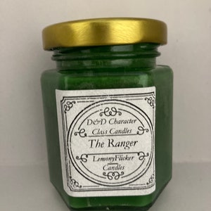 Puede incluir: Una vela de vidrio verde en un frasco con tapa dorada. La etiqueta dice "D&D Character Class Candles The Ranger Lemony Flicker Candles".