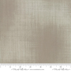 May include: A neutral-toned fabric with a textured appearance. The fabric has a subtle, mottled effect, with vertical and horizontal lines creating a woven look. The bottom of the image shows a ruler with inch measurements. The fabric is suitable for quilting and sewing projects.
