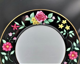 May include: Black and white decorative plate with a floral design. The plate features a black rim with a gold border and is adorned with pink and yellow flowers and green leaves.