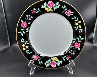 May include: Black dinner plate with a gold rim and a floral design featuring pink, yellow, and green flowers.