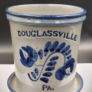 May include: A white ceramic crock with a blue floral design and the words "DOUGLASSVILLE PA."  The crock has a matching saucer.