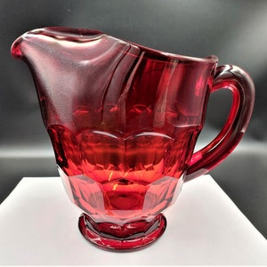 May include: A red glass pitcher with a faceted design and a handle. The pitcher has a rounded base and a wide mouth.