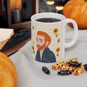 Può includere: Una tazza in ceramica bianca con un'illustrazione grafica di una persona con capelli rossi e barba, che indossa una giacca blu scuro a motivi. Il design include girasoli gialli, piccole foglie verdi e stelline su uno sfondo beige chiaro.