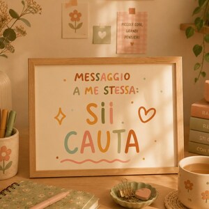 Puede incluir: Un marco de madera rectangular que contiene una impresión blanca con el texto en italiano MESSAGGIO A ME STESSA: Sii CAUTA en letras multicolores. El diseño incluye una pequeña estrella naranja, un corazón y una línea ondulada sobre fondo liso.