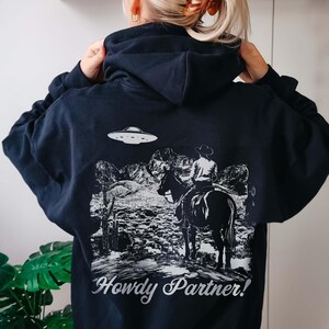 May include: Black hoodie with a white graphic print of a cowboy riding a horse with a UFO in the background. The text "Howdy Partner!" is printed below the graphic.