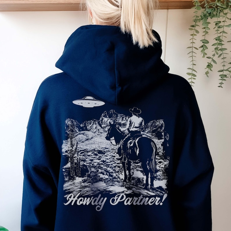 May include: A navy blue hoodie with a white graphic print of a cowboy riding a horse in front of a mountain range. A UFO is flying above the cowboy. The text "Howdy Partner!" is printed below the graphic.