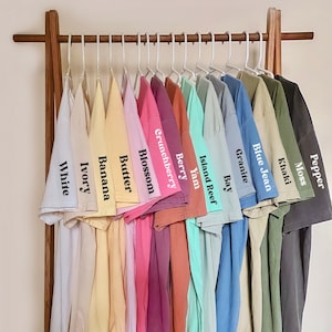 Puede incluir: Un estante con 14 camisetas de diferentes colores, colgadas en perchas. Las camisetas est&aacute;n etiquetadas con nombres como "White", "Ivory", "Banana", "Butter", "Blossom", "Crunchberry", "Berry", "Yam", "Island Reef", "Bay", "Granite", "Blue Jean", "Khaki" y "Moss".