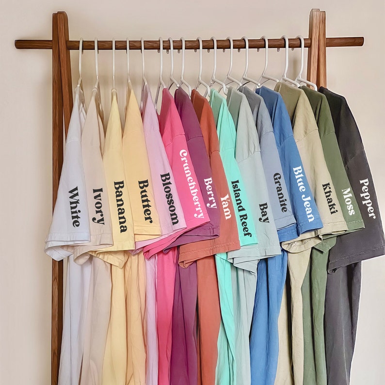 Puede incluir: Un estante con 14 camisetas de diferentes colores, colgadas en perchas. Las camisetas est&aacute;n etiquetadas con nombres como "White", "Ivory", "Banana", "Butter", "Blossom", "Crunchberry", "Berry", "Yam", "Island Reef", "Bay", "Granite", "Blue Jean", "Khaki" y "Moss".