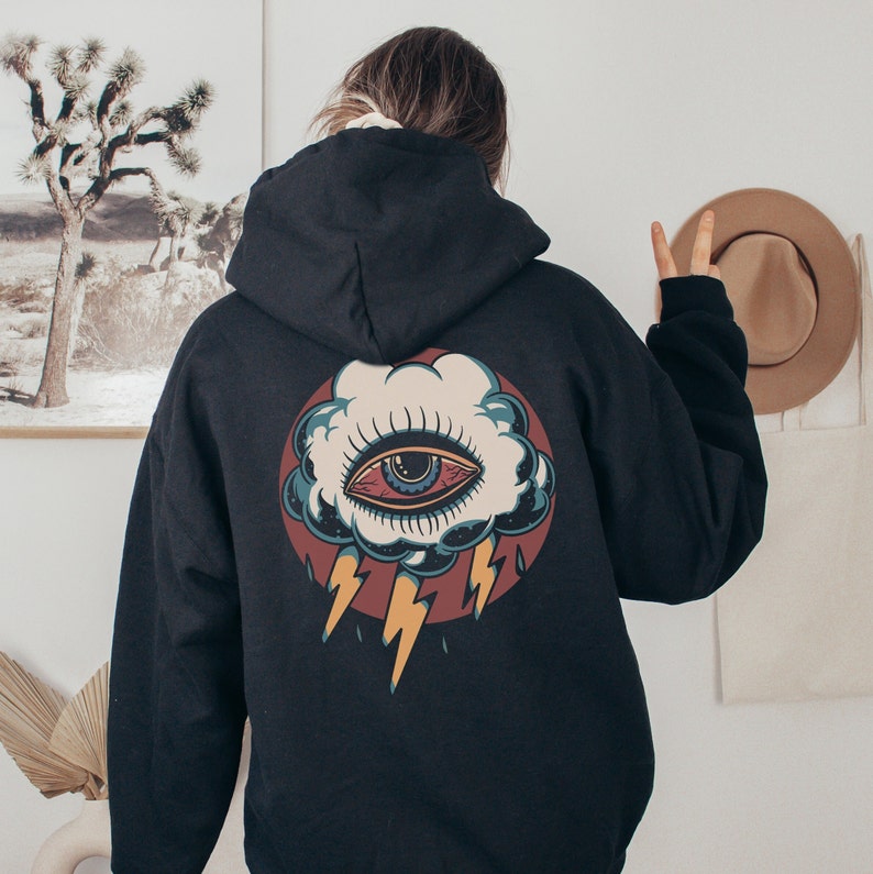Op de afbeelding: Zwart hoodie met een grafische print van een gestileerd oog in een wolk met bliksem. Het oog is blauw met rode en witte accenten.