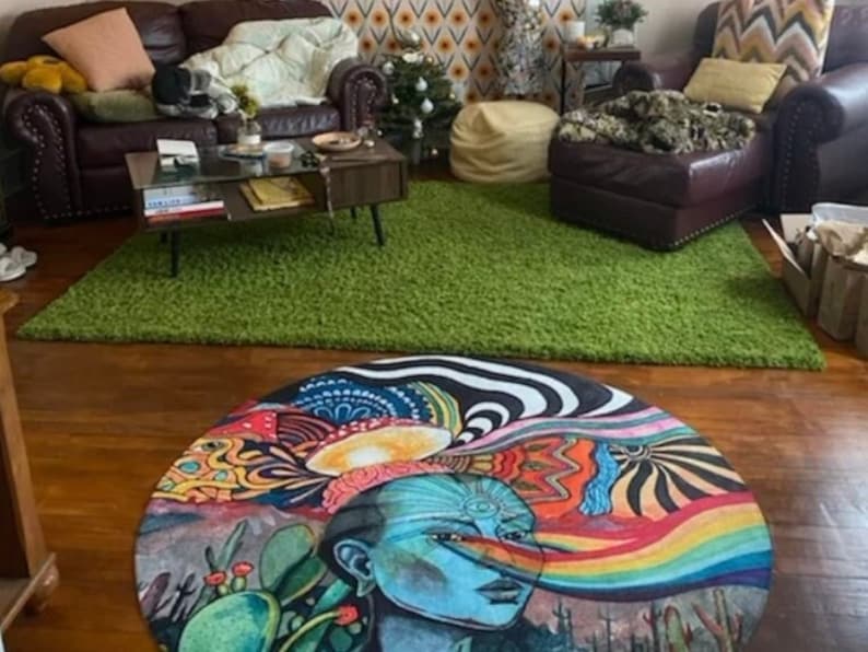 Pu&ograve; includere: Un tappeto rotondo e colorato con un design psichedelico che presenta un viso con un terzo occhio e un arcobaleno. Il tappeto &egrave; su un pavimento di legno con un tappeto di pelo verde sullo sfondo.