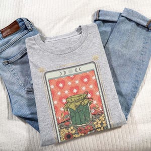 May include: A gray t-shirt with a graphic of a tarot card featuring a jar of pickles, hearts, stars, and flowers. The card is titled "The Pickle Card".