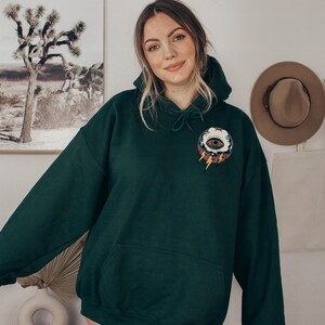 Op de afbeelding: Een donkergroen hoodie met een zak op de voorkant. De hoodie heeft een afbeelding van een oog in een wolk met bliksemstralen op de voorkant.