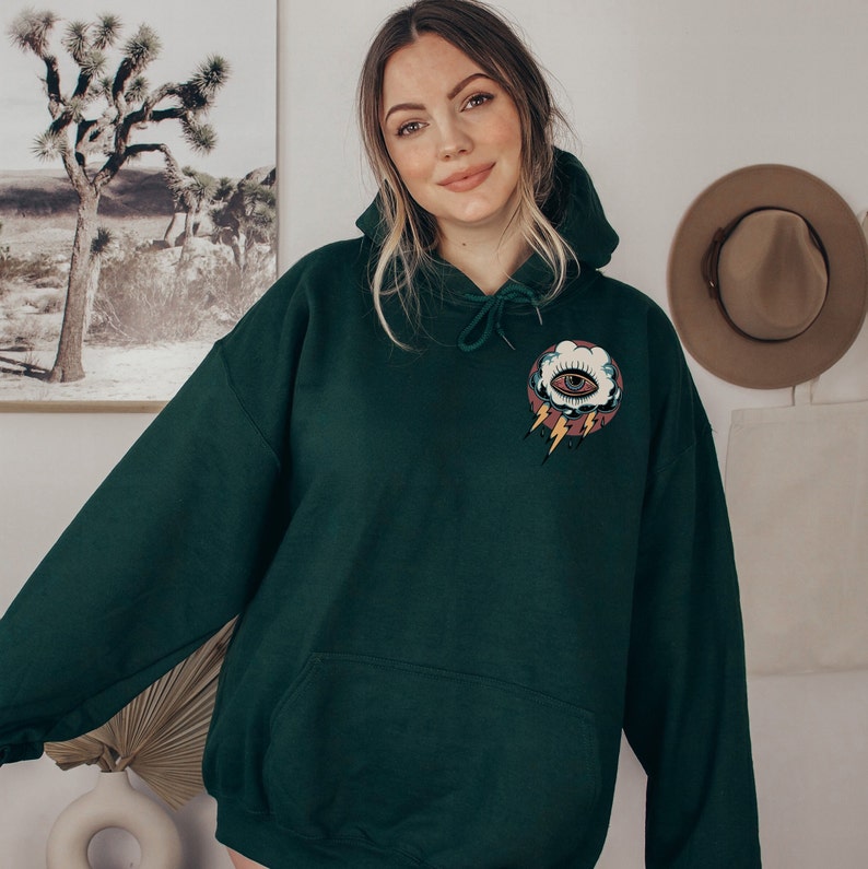 Op de afbeelding: Een donkergroen hoodie met een zak op de voorkant. De hoodie heeft een afbeelding van een oog in een wolk met bliksemstralen op de voorkant.