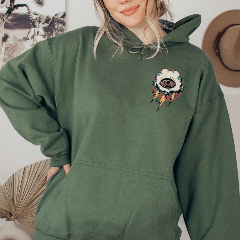 Op de afbeelding: Een donkergroen hoodie met een zak op de voorkant. De hoodie heeft een grafische print van een gestileerd oog in een wolk met bliksem en regendruppels.
