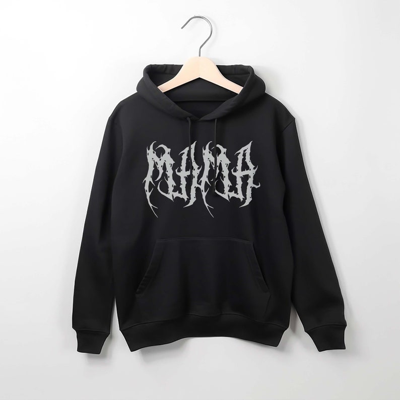 Puede incluir: Sudadera con capucha negra con un bolsillo delantero y una capucha, colgada de una percha de madera. La sudadera presenta la palabra "MUMMA" en plata, con una fuente de estilo g&oacute;tico en el pecho.