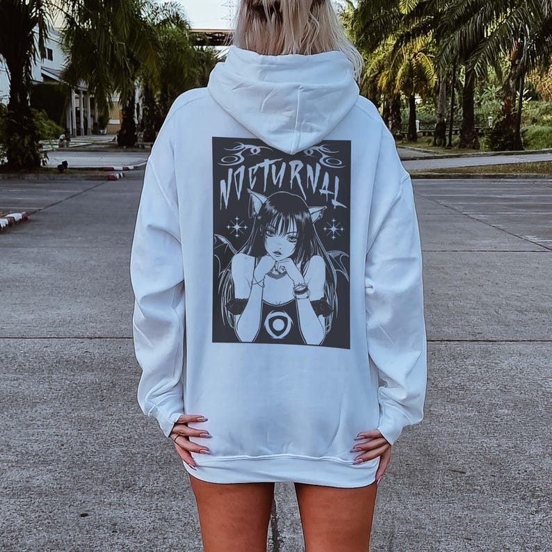 Pu&ograve; includere: Una felpa con cappuccio bianca con una grafica in bianco e nero sul retro. La grafica &egrave; un'illustrazione di una donna con orecchie da gatto e la scritta "NOCTURNAL" sopra. La felpa ha maniche lunghe e una tasca a marsupio.