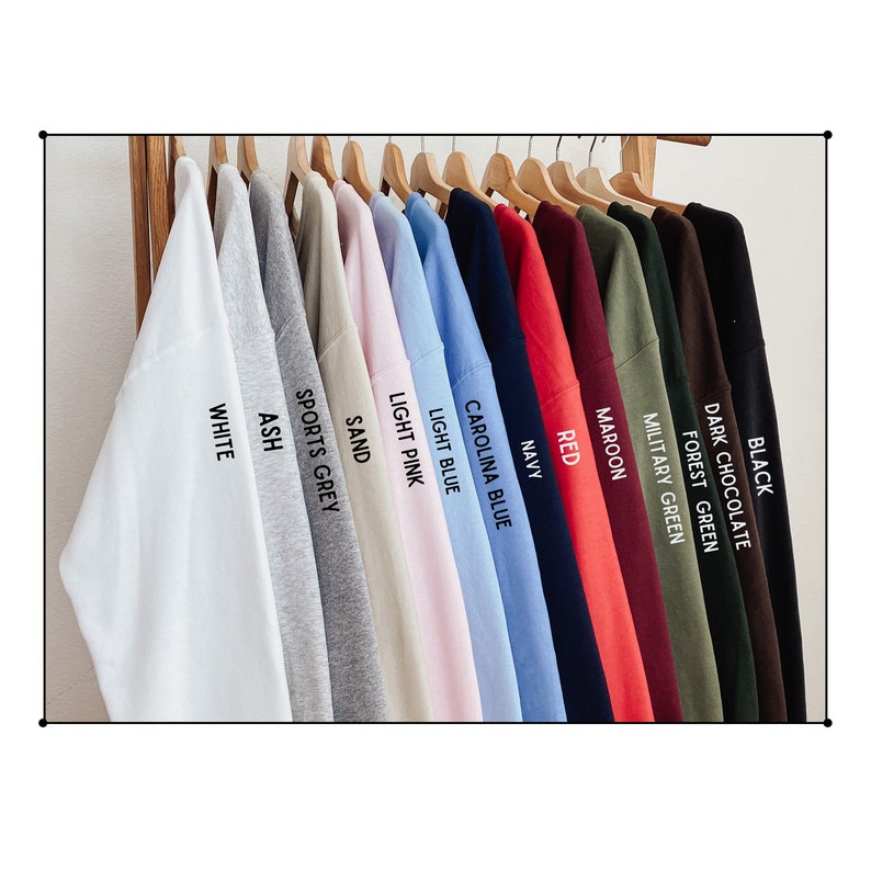 Puede incluir: Una fila de camisetas colgadas en un estante de madera, mostrando una variedad de colores, incluyendo blanco, ceniza, gris deportivo, arena, rosa claro, azul claro, azul carolina, azul marino, rojo, granate, verde militar, verde bosque, chocolate negro y negro.