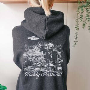May include: A dark gray hoodie with a white graphic print of a cowboy riding a horse in front of a mountain range with a UFO in the sky. The text "Howdy Partner!" is printed below the graphic.