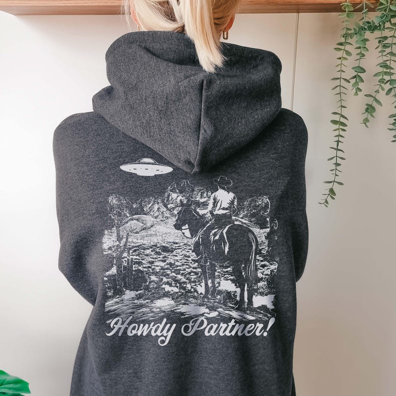 May include: A dark gray hoodie with a white graphic print of a cowboy riding a horse in front of a mountain range with a UFO in the sky. The text "Howdy Partner!" is printed below the graphic.