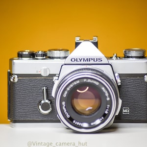 Puede incluir: Una cámara Olympus OM-System vintage, con cuerpo plateado y negro, lente prominente y el logotipo de Olympus. La cámara se encuentra sobre un fondo amarillo vibrante, mostrando su diseño clásico y detalles.