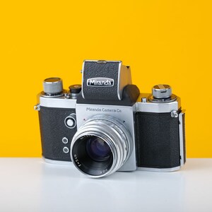 May include: A silver and black Miranda Camera Co. 35mm film camera with a silver lens. The camera has a black leatherette covering and a silver top plate with the Miranda logo.