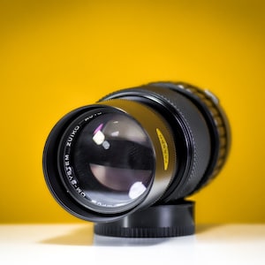 Peut inclure: Objectif d'appareil photo Olympus OM-SYSTEM Zuiko Auto noir avec un pare-soleil. L'objectif est incliné, le devant face au spectateur. L'objectif est posé sur un fond jaune vif.