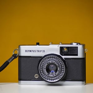 Puede incluir: Una c&aacute;mara de pel&iacute;cula Olympus Trip 35 plateada y negra con una correa negra. La c&aacute;mara tiene una lente negra con un anillo plateado.
