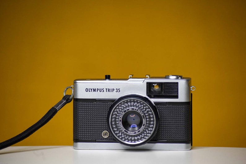 Puede incluir: Una c&aacute;mara de pel&iacute;cula Olympus Trip 35 plateada y negra con una correa negra. La c&aacute;mara tiene una lente negra con un anillo plateado.