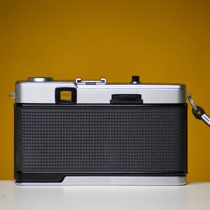 Puede incluir: Una c&aacute;mara de pel&iacute;cula de 35 mm plateada y negra con una correa negra. La c&aacute;mara tiene un cuerpo negro texturizado y una lente plateada.