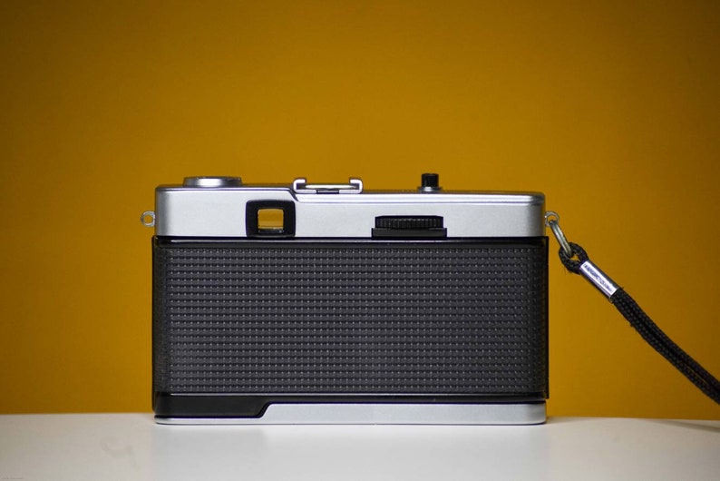 Puede incluir: Una c&aacute;mara de pel&iacute;cula de 35 mm plateada y negra con una correa negra. La c&aacute;mara tiene un cuerpo negro texturizado y una lente plateada.