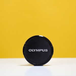 Puede incluir: Una tapa de lente de cámara negra con la palabra "OLYMPUS" impresa en blanco en la parte delantera.