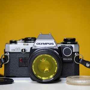 May include: An Olympus OM-10 film camera with a Zuiko Auto-S 50mm lens. The camera is silver and black, with a black leather strap. A lens cap and a clear plastic lens protector are in front of the camera. The background is a solid yellow.