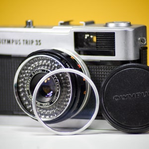 Op de afbeelding: Een vintage Olympus Trip 35 camera, zilver en zwart, met een lens, filter en lensdop. De camera heeft de tekst "OLYMPUS TRIP 35". De lens heeft een zilveren rand en de lensdop is zwart.