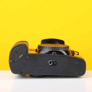 Puede incluir: C&aacute;mara de pel&iacute;cula negra con un dial plateado en la parte superior. La c&aacute;mara est&aacute; sobre una superficie amarilla.