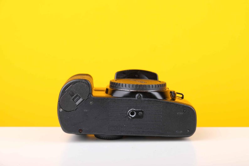 Puede incluir: C&aacute;mara de pel&iacute;cula negra con un dial plateado en la parte superior. La c&aacute;mara est&aacute; sobre una superficie amarilla.