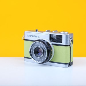 May include: An Olympus Trip 35 camera with a silver body and a light green leatherette grip. The camera lens is black and silver, and the background is a bright yellow. The camera is on a white surface.