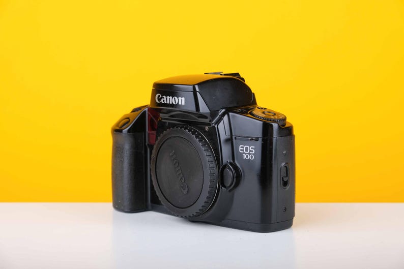 Puede incluir: Una c&aacute;mara de pel&iacute;cula Canon EOS 100 negra con una tapa de lente en el lente. La c&aacute;mara est&aacute; sobre una superficie blanca con un fondo amarillo.