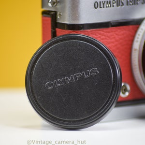 Op de afbeelding: Close-up van een zwarte Olympus lensdop met de merknaam in reliëf. De dop bedekt gedeeltelijk een vintage Olympus Trip 35 camera met een rode leren behuizing en zilveren accenten. De achtergrond is heldergeel.